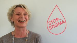 A woman smiling widely at the camera, with short blonde hair and a grey jumper. A logo saying stop the stigma is on the right
